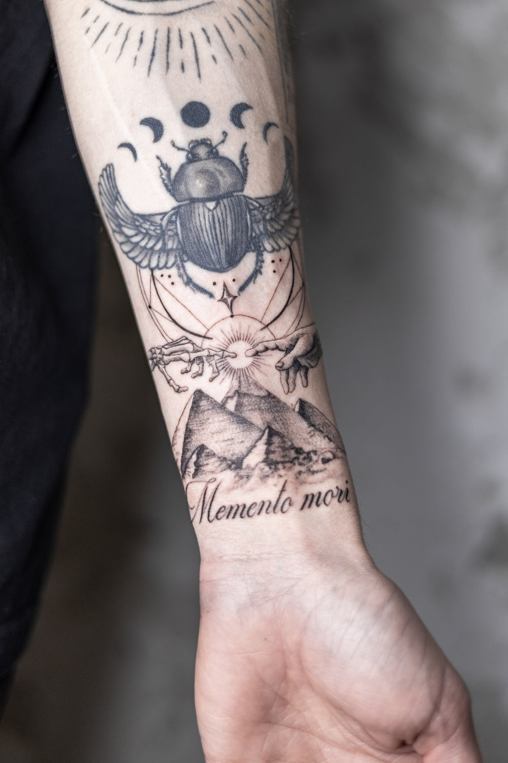 Tattooed arm with a beetle, pyramid, and 'Memento mori' text on a blurred background
