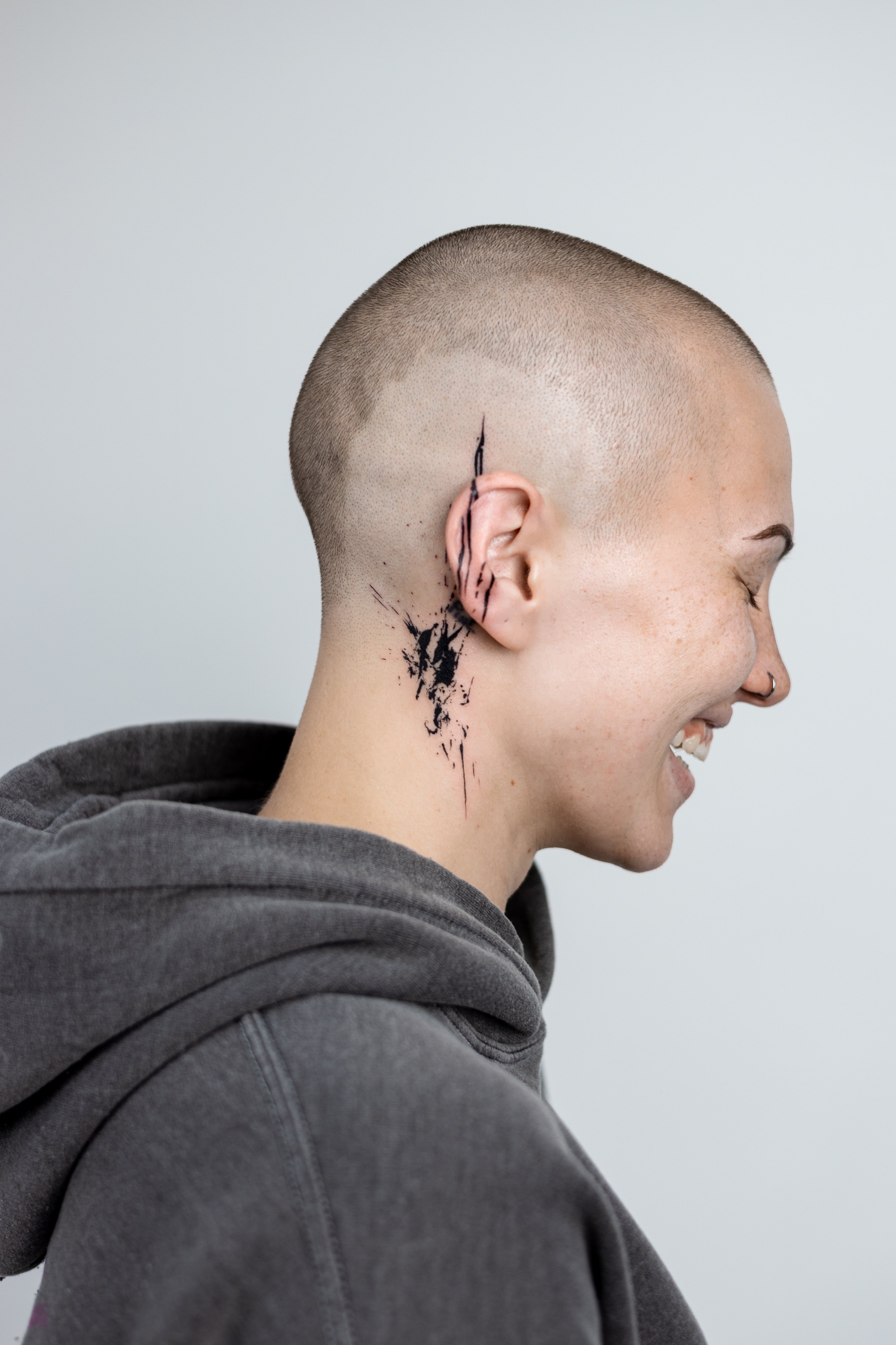 Person with a shaved head and visible neck tattoo wearing a gray hoodie against a light gray background